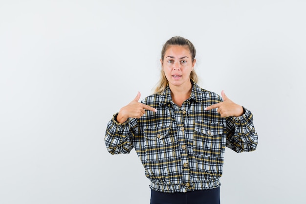 Señorita señalando a sí misma en camisa, pantalones cortos y mirando confiada, vista frontal.