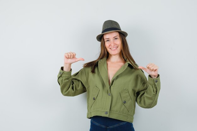 Señorita señalando a sí misma con los pulgares en el sombrero de los pantalones de la chaqueta y mirando orgullosa