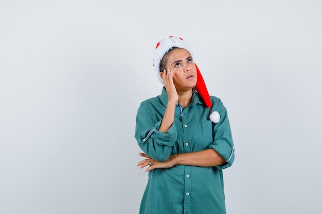 Señorita con sombrero de navidad, camisa con la mano cerca de la cara y mirando pensativo, vista frontal.