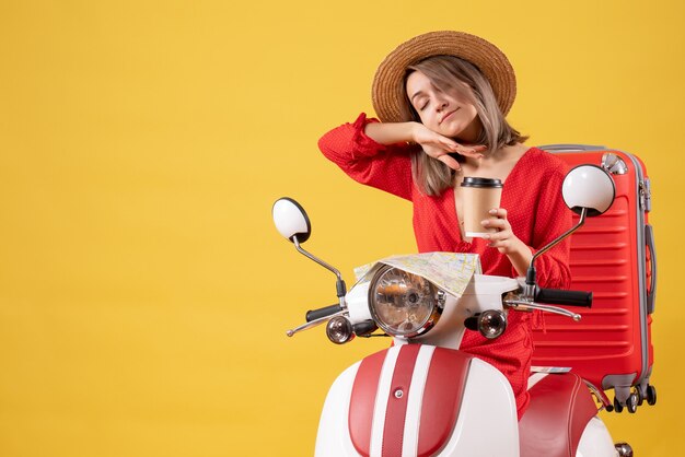 Señorita soñolienta en vestido rojo sosteniendo una taza de café cerca del ciclomotor