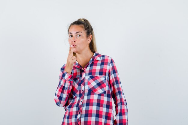 Señorita soplando pistola de dedo en camisa a cuadros y mirando confiado. vista frontal.