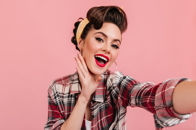 Señorita sorprendida en traje retro mirando a la cámara. Encantadora chica pinup tomando selfie sobre fondo rosa.