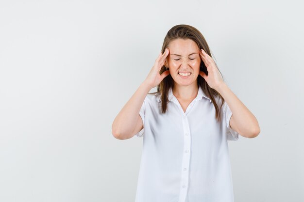 Señorita sosteniendo la cabeza en una blusa blanca y luciendo incómoda