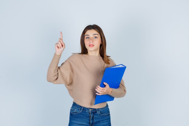 Señorita sosteniendo una carpeta azul mientras apunta hacia arriba en suéter beige, jeans y mirando con cuidado. vista frontal.