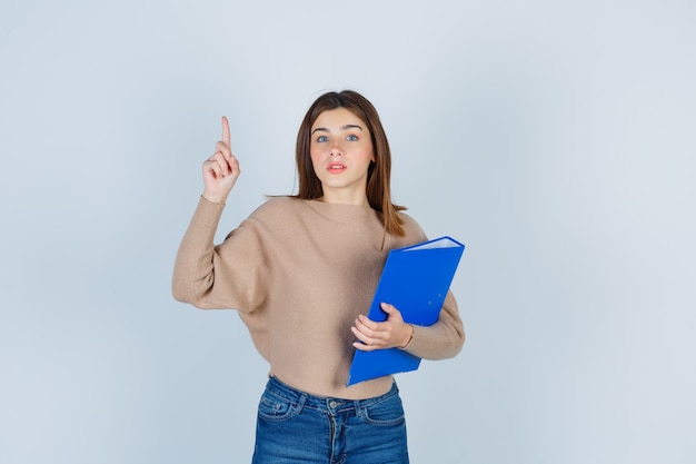 Señorita sosteniendo una carpeta azul mientras apunta hacia arriba en suéter beige, jeans y mirando con cuidado. vista frontal.