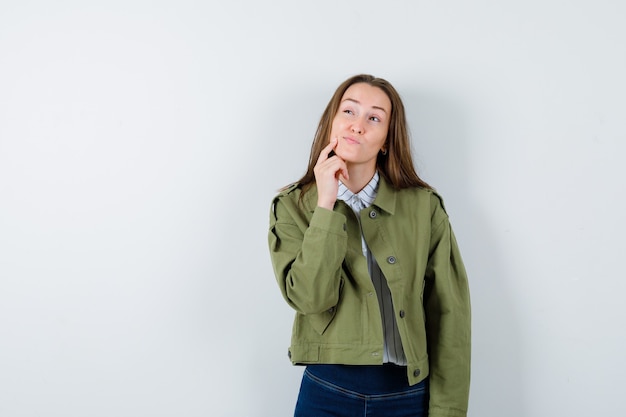 Señorita sosteniendo el dedo en la barbilla en blusa, chaqueta y mirando pensativo.