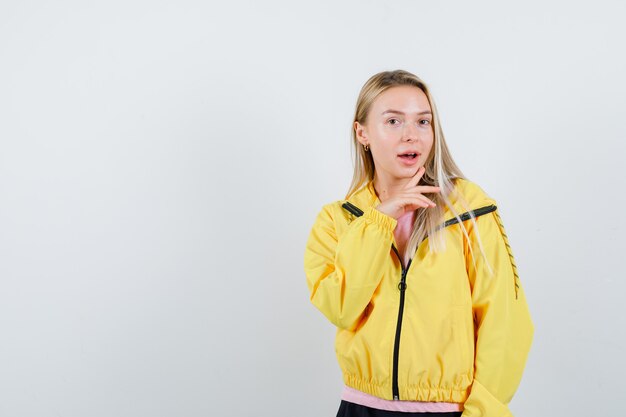 Señorita sosteniendo el dedo debajo de la barbilla en camiseta, chaqueta y mirando asombrado