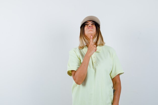 Señorita sosteniendo el dedo debajo de la barbilla mientras mira hacia arriba en camiseta, gorra y mirando pensativo, vista frontal.