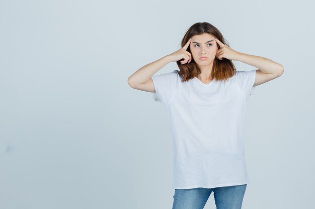 Señorita sosteniendo los dedos en la cabeza en camiseta, jeans y mirando pensativo. vista frontal.