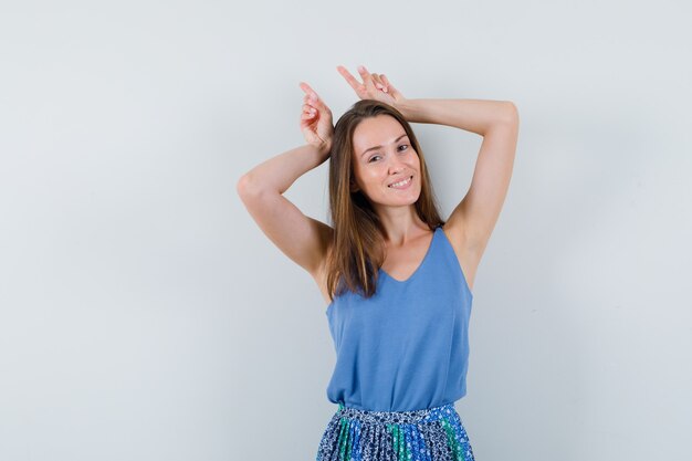 Señorita sosteniendo los dedos sobre la cabeza como cuernos de toro en camiseta, falda y aspecto divertido