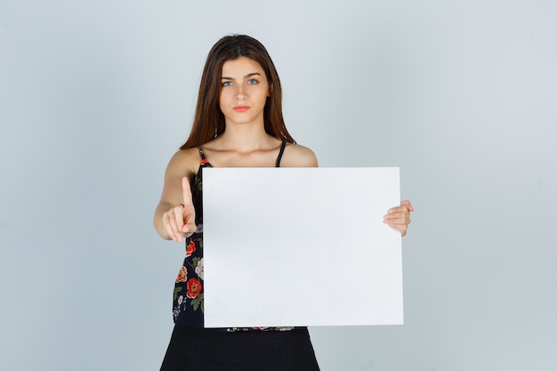 Foto gratuita señorita sosteniendo un lienzo en blanco, mostrando mantener un gesto de un minuto en blusa, falda y mirando serio, vista frontal.