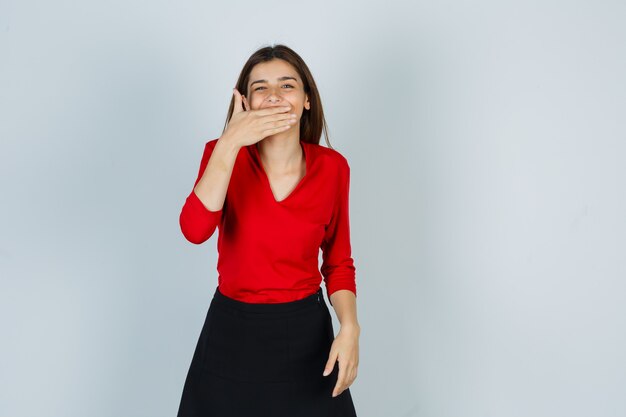 Señorita sosteniendo la mano en la boca en blusa roja, falda y luciendo linda