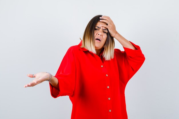 Señorita sosteniendo la mano en la cabeza mientras extiende la palma hacia un lado en una camisa roja de gran tamaño y mirando ansiosa, vista frontal.