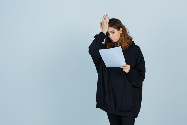 Señorita sosteniendo la mano en la cabeza mientras mantiene el papel en una sudadera con capucha de gran tamaño, pantalones y mirando olvidadizo, vista frontal.