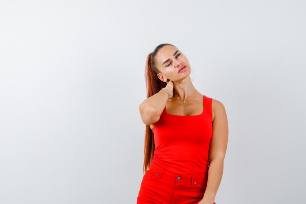 Señorita sosteniendo la mano detrás del cuello en camiseta roja, pantalón rojo y aspecto cansado. vista frontal.