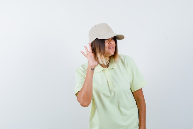 Señorita sosteniendo la mano detrás de la oreja en camiseta, gorra y mirando enfocado. vista frontal.