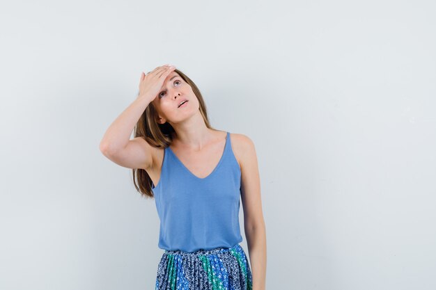 Señorita sosteniendo la mano en la frente con blusa azul, falda y aspecto cansado, vista frontal.