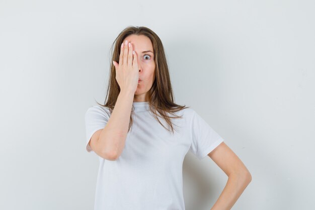 Señorita sosteniendo la mano en un ojo en camiseta blanca y mirando sorprendido