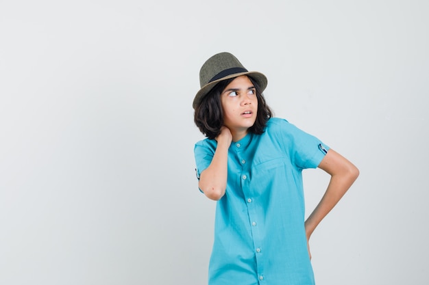 Señorita sosteniendo la mano en su cuello mientras mira a otro lado con camisa azul, sombrero y parece asustada.