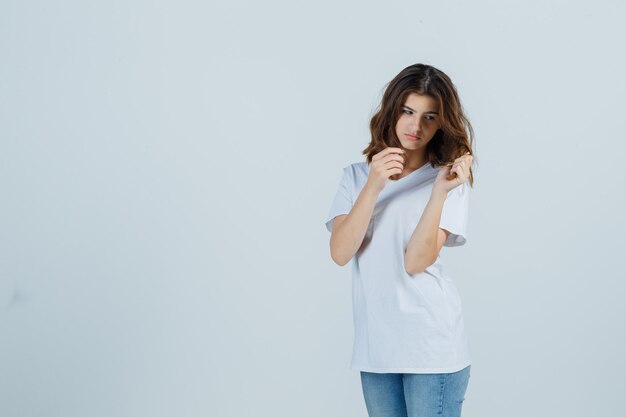 Señorita sosteniendo un mechón de cabello en camiseta, jeans y mirando pensativo. vista frontal.