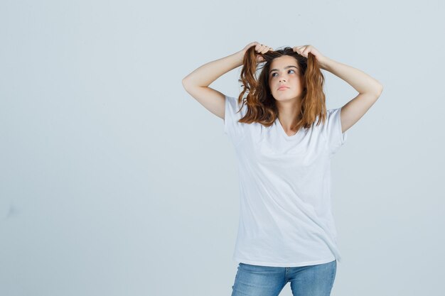 Señorita sosteniendo un mechón de cabello en camiseta, jeans y mirando pensativo. vista frontal.