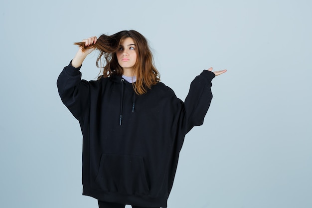 Señorita sosteniendo un mechón de cabello en una sudadera con capucha de gran tamaño, pantalones y mirando pensativo, vista frontal.