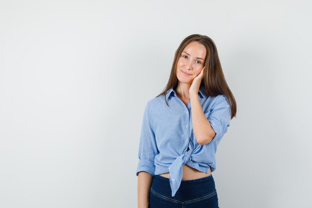 Señorita sosteniendo la palma en la mejilla con camisa azul, pantalones y aspecto lindo