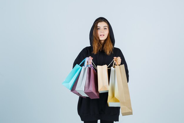 Señorita sosteniendo paquetes mientras mira a la cámara en una sudadera con capucha de gran tamaño, pantalones y mirando asombrado, vista frontal.