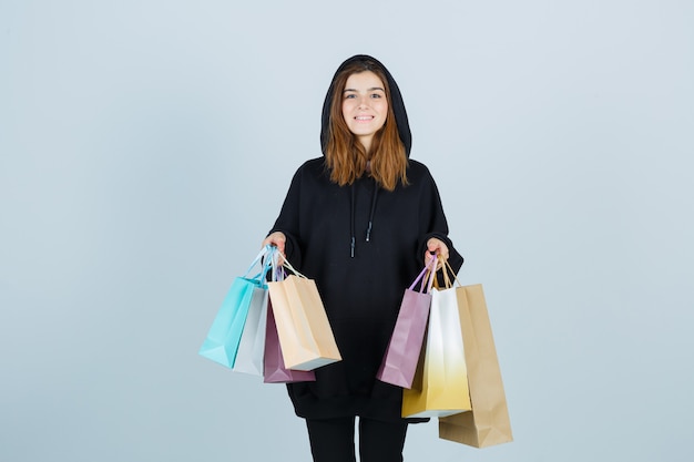 Señorita sosteniendo paquetes mientras mira a la cámara en una sudadera con capucha de gran tamaño, pantalones y parece feliz. vista frontal.