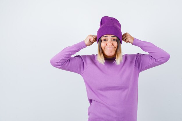 Señorita sosteniendo puños en la cabeza en suéter morado, gorro y mirando divertido. vista frontal.