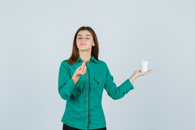 Señorita sosteniendo una taza de café de plástico mientras muestra un gesto delicioso en camisa y parece complacido, vista frontal.