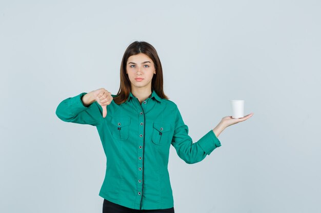 Señorita sosteniendo una taza de café de plástico mientras muestra el pulgar hacia abajo en la camisa y mira insatisfecho, vista frontal.