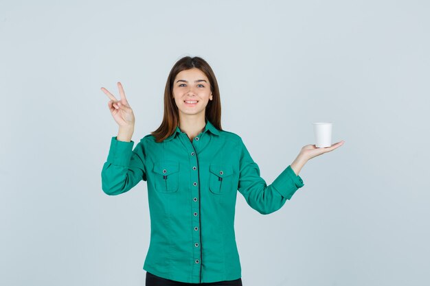 Señorita sosteniendo una taza de café de plástico mientras muestra el signo de la victoria en camisa y se ve alegre. vista frontal.
