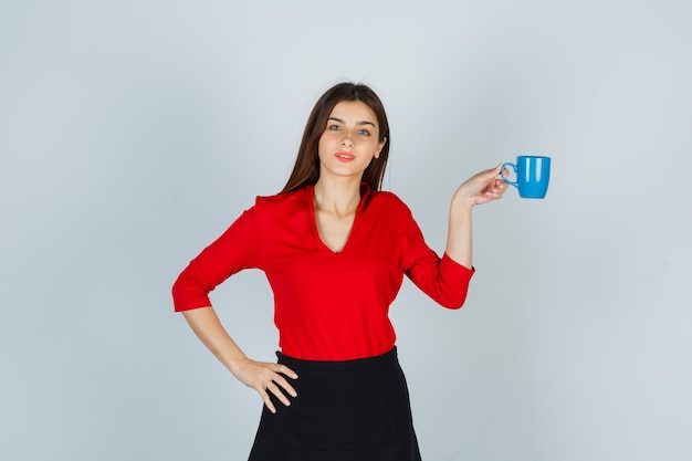 Señorita sosteniendo la taza mientras mantiene la mano en la cadera en blusa roja