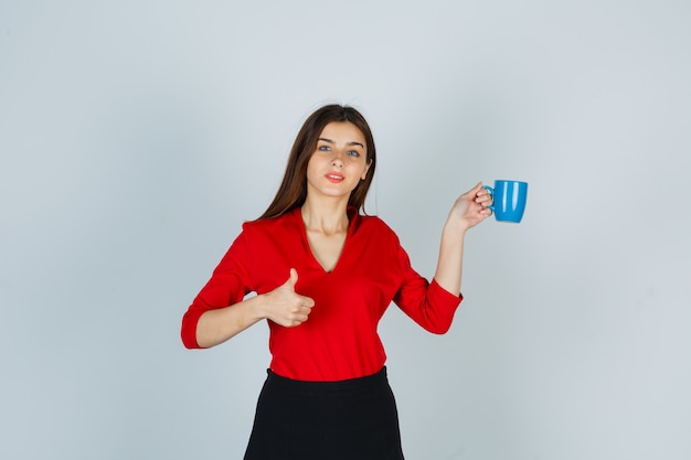 Foto gratuita señorita sosteniendo la taza mientras muestra el pulgar hacia arriba en blusa roja, falda y parece confiada