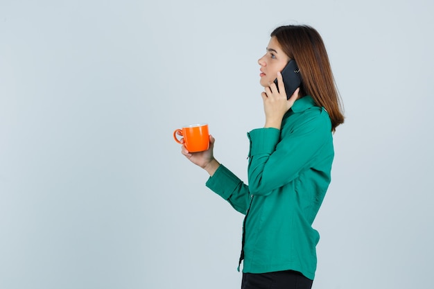 Señorita sosteniendo una taza de té naranja, hablando por el teléfono móvil en camisa y mirando pensativo, vista frontal.