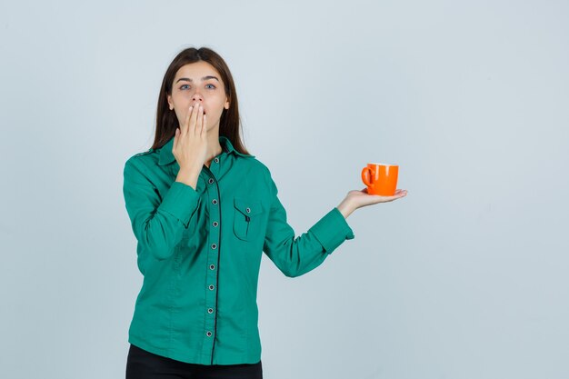 Señorita sosteniendo una taza de té naranja, manteniendo la mano en la boca en la camisa y mirando enfocado, vista frontal.