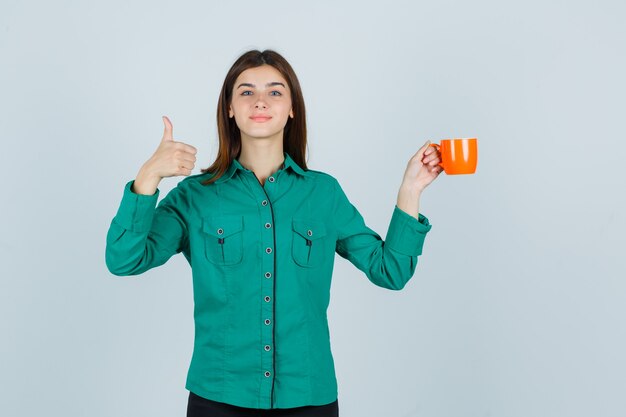 Señorita sosteniendo una taza de té naranja mientras muestra el pulgar hacia arriba en la camisa y parece complacido. vista frontal.