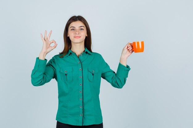 Señorita sosteniendo una taza de té naranja mientras muestra el signo de ok en camisa y parece seguro de sí mismo. vista frontal.