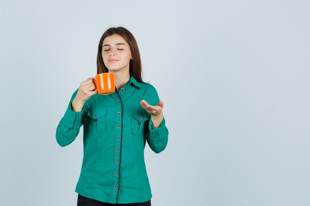 Señorita sosteniendo una taza de té naranja, mostrando algo en camisa y luciendo pacífica. vista frontal.