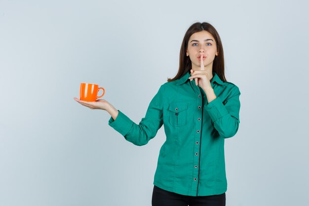 Señorita sosteniendo una taza de té naranja, mostrando gesto de silencio en camisa y mirando con cuidado. vista frontal.