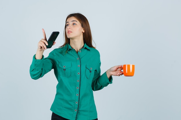 Señorita sosteniendo una taza de té naranja y un teléfono móvil, apuntando hacia afuera en camisa y mirando con cuidado. vista frontal.