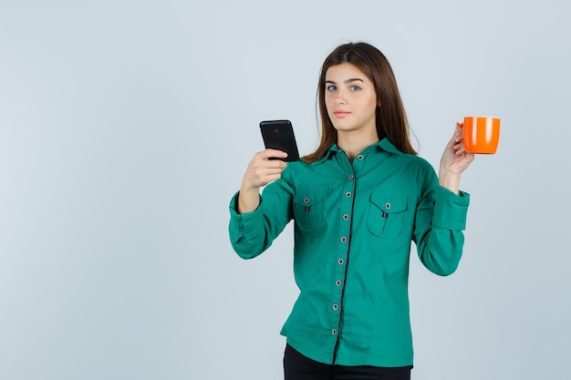 Señorita sosteniendo una taza de té naranja y un teléfono móvil en camisa y mirando confiado, vista frontal.