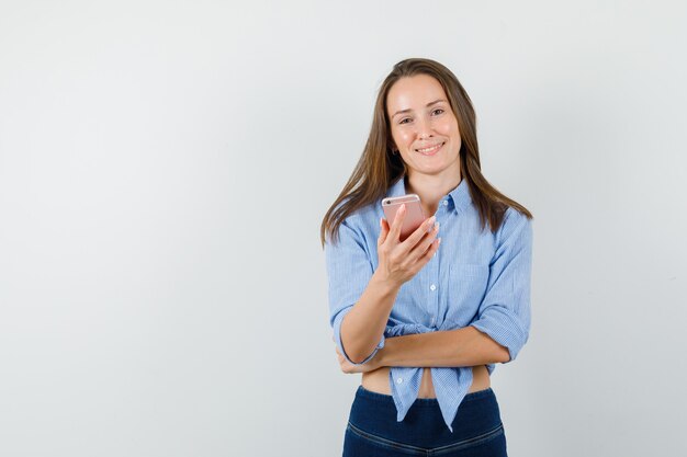 Señorita sosteniendo teléfono móvil en camisa azul, pantalones y mirando alegre