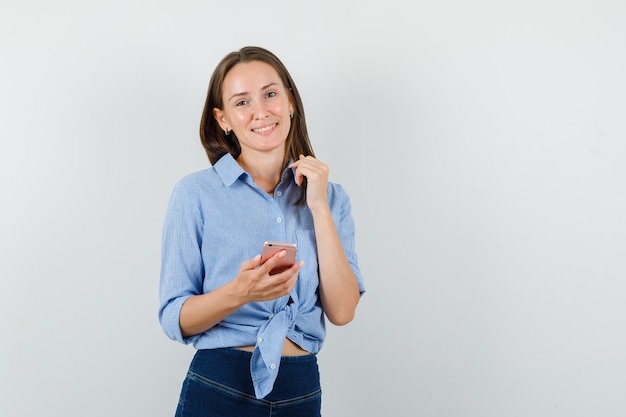 Señorita sosteniendo teléfono móvil en camisa azul, pantalones y mirando alegre