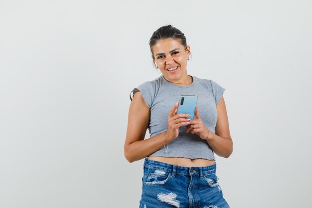 Señorita sosteniendo teléfono móvil en camiseta, pantalones cortos y mirando alegre.