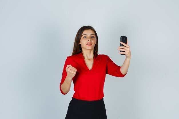Señorita sosteniendo teléfono móvil mientras levanta el puño en blusa roja