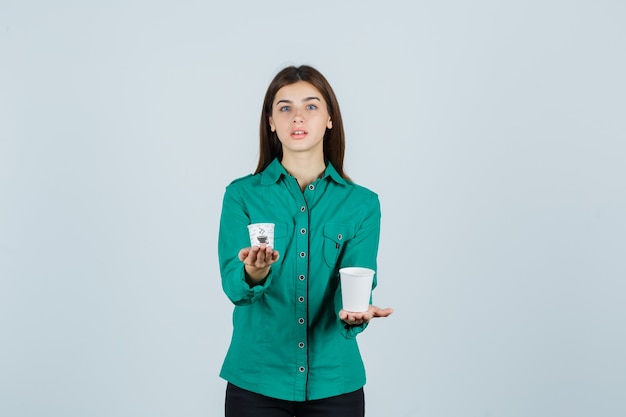 Señorita sosteniendo vasos de plástico de café en camisa y mirando confiado, vista frontal.