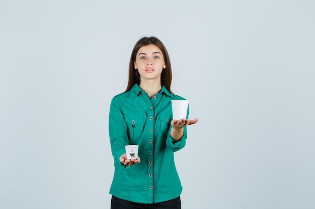 Señorita sosteniendo vasos de plástico de café en camisa y mirando confiado, vista frontal.