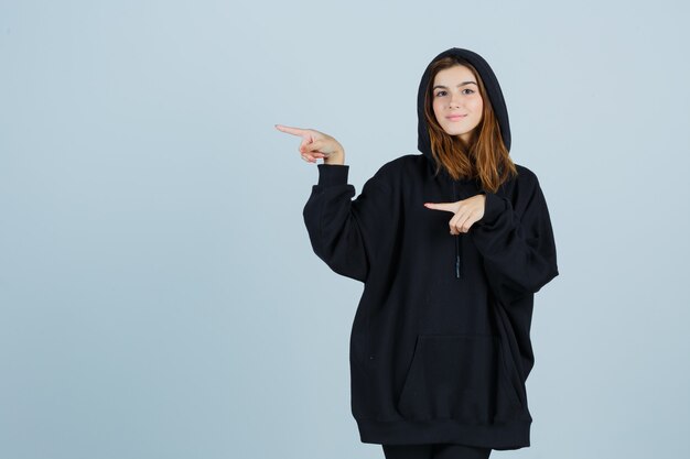 Señorita en sudadera con capucha de gran tamaño, pantalones apuntando hacia el lado izquierdo y mirando confiado, vista frontal.